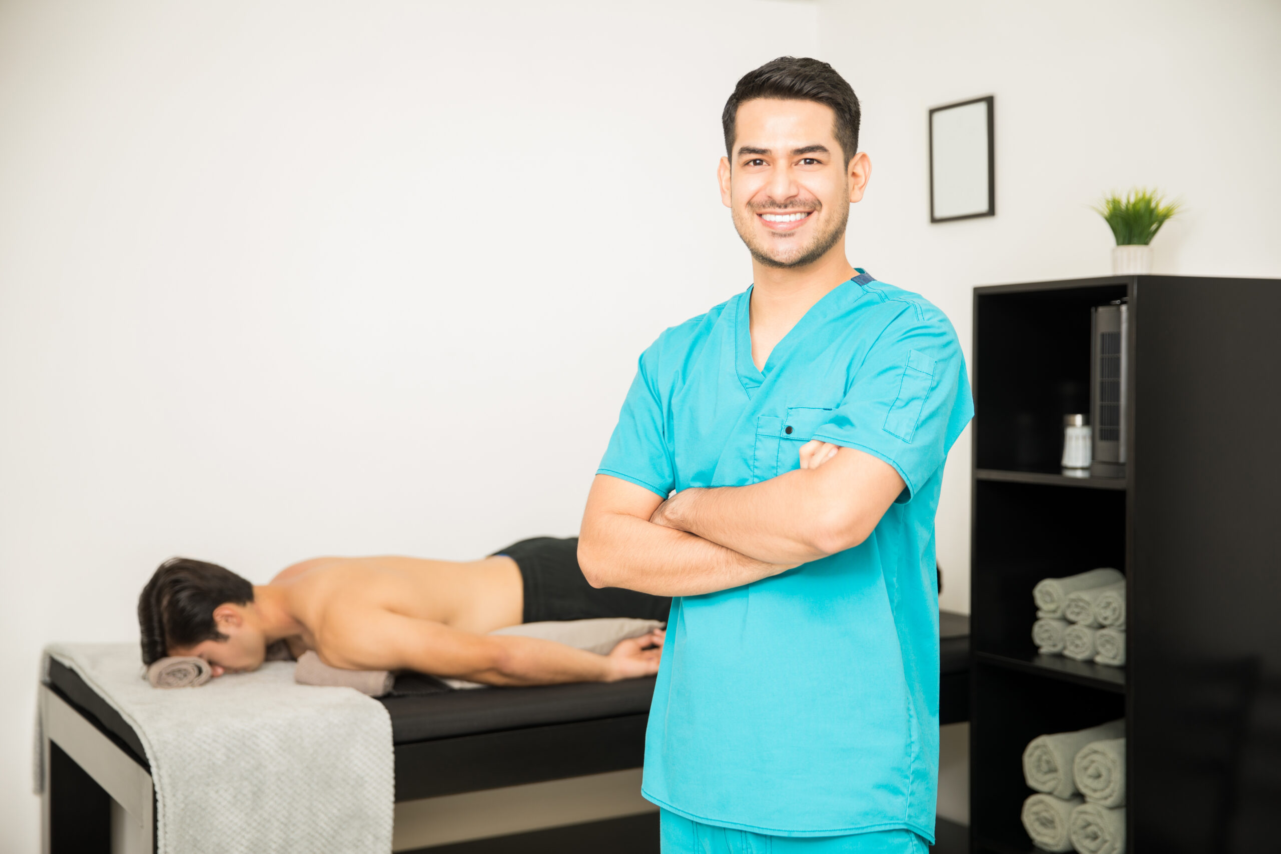 Smiling male physiotherapist standing arms crossed with patient lying in background at clinic